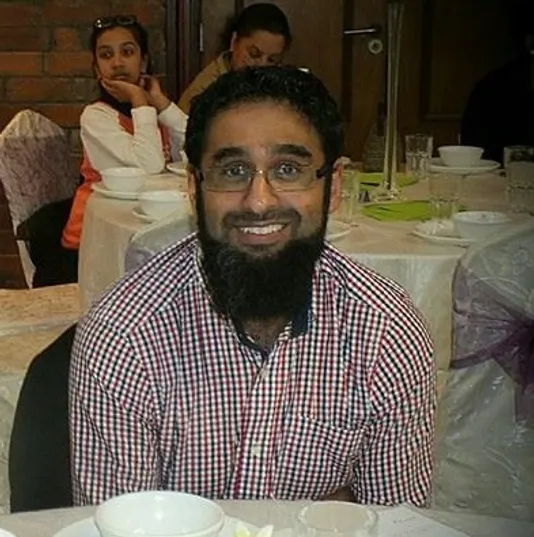 a picture of a bearded man in a white and red small checkered t-shirt smiling at the camera, with a round table behind him with plates and a lady seated at the table